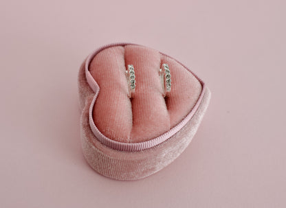 A pair of kids hypoallergenic silver earrings with small clear stones, displayed in a pink velvet heart-shaped ring box on a solid pink background. The earrings are from the brand The Pierced Peanut Earring Co in New Zealand, NZ.