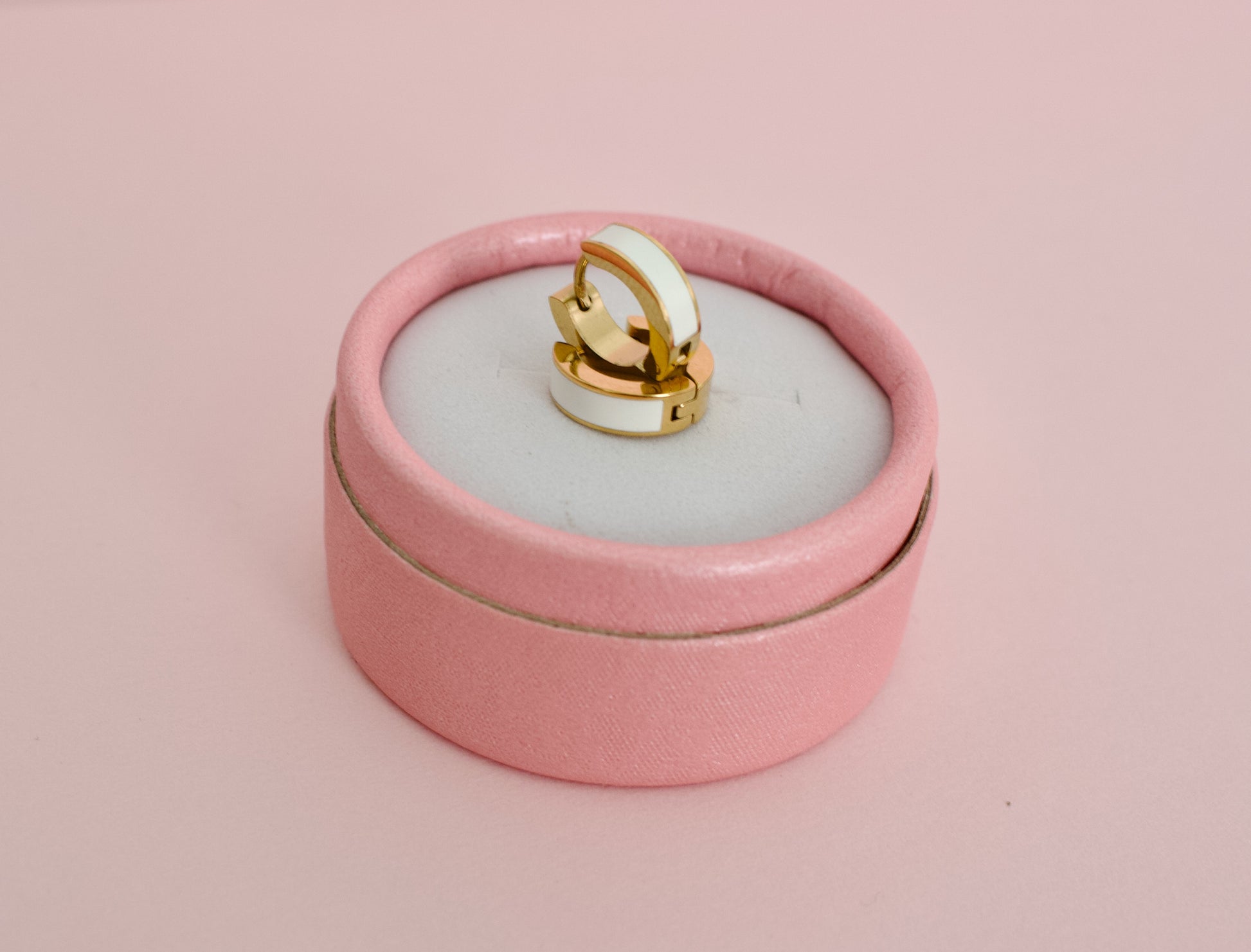 A pair of kids hypoallergenic gold huggie hoop earrings with a white enamel inlay, displayed on a round pink velvet ring box against a solid pink background from the brand The Pierced Peanut Earring Co in New Zealand, NZ.