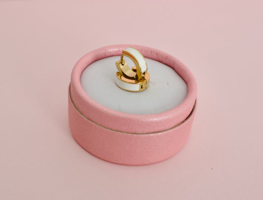 A pair of kids hypoallergenic gold huggie hoop earrings with a white enamel inlay, displayed on a round pink velvet ring box against a solid pink background from the brand The Pierced Peanut Earring Co in New Zealand, NZ.
