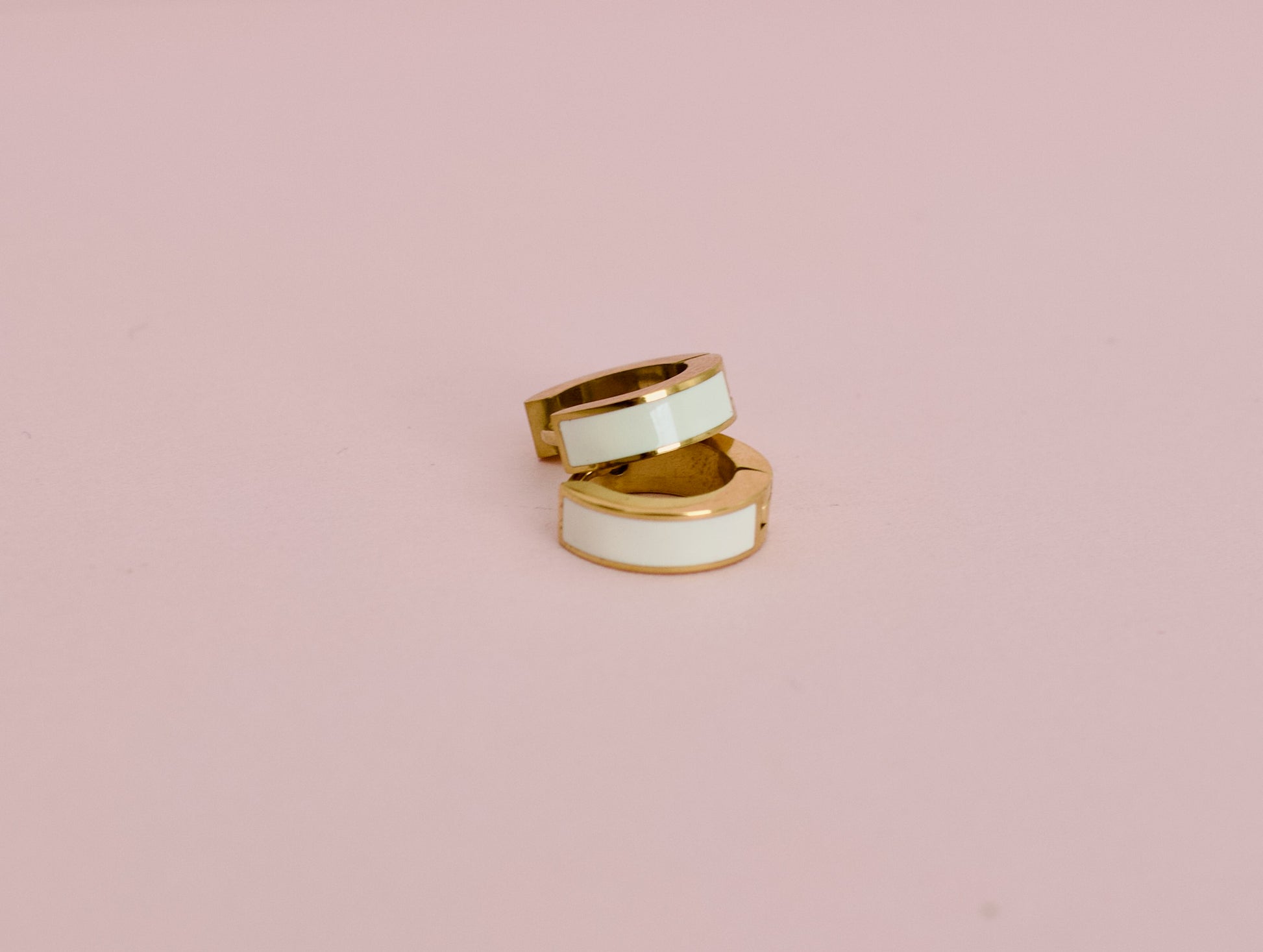 A pair of kids hypoallergenic gold huggie hoop earrings with a white enamel inlay, displayed against a solid pink background from the brand The Pierced Peanut Earring Co in New Zealand, NZ.