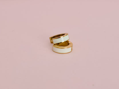 A pair of kids hypoallergenic gold huggie hoop earrings with a white enamel inlay, displayed against a solid pink background from the brand The Pierced Peanut Earring Co in New Zealand, NZ.