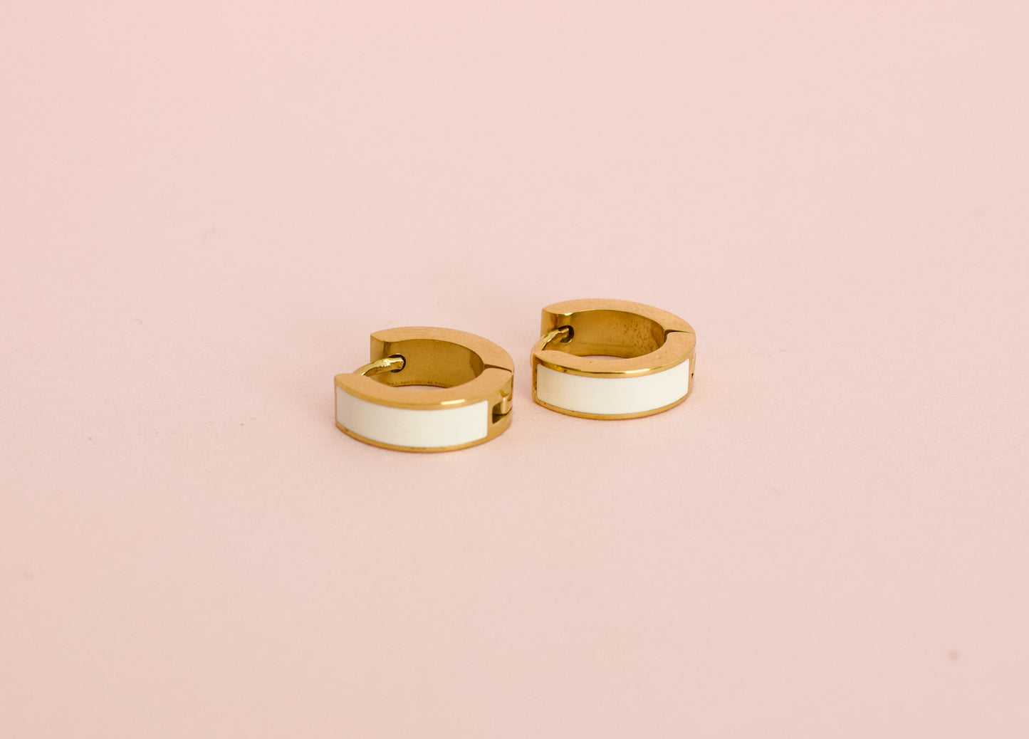 A pair of kids hypoallergenic gold huggie hoop earrings with a white enamel inlay, displayed against a solid pink background from the brand The Pierced Peanut Earring Co in New Zealand, NZ.