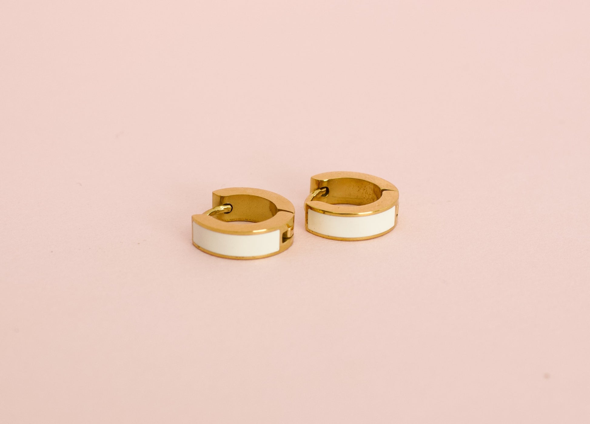 A pair of kids hypoallergenic gold huggie hoop earrings with a white enamel inlay, displayed against a solid pink background from the brand The Pierced Peanut Earring Co in New Zealand, NZ.