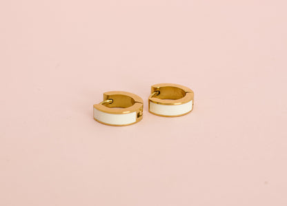 A pair of kids hypoallergenic gold huggie hoop earrings with a white enamel inlay, displayed against a solid pink background from the brand The Pierced Peanut Earring Co in New Zealand, NZ.