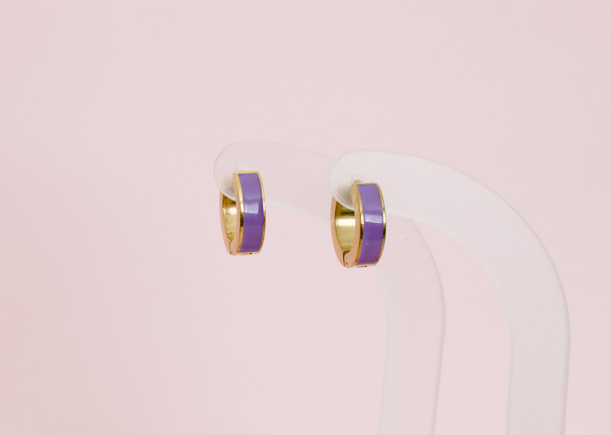 A pair of kids small hypoallergenic gold hoop earrings with a smooth purple enamel inlay, displayed on a clear acrylic stand against a solid pale pink background. The earrings are from the brand The Pierced Peanut Earring Co in New Zealand, NZ.