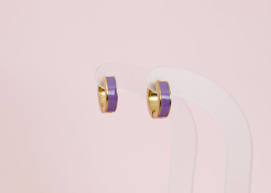 A pair of kids small hypoallergenic gold hoop earrings with a smooth purple enamel inlay, displayed on a clear acrylic stand against a solid pale pink background. The earrings are from the brand The Pierced Peanut Earring Co in New Zealand, NZ.