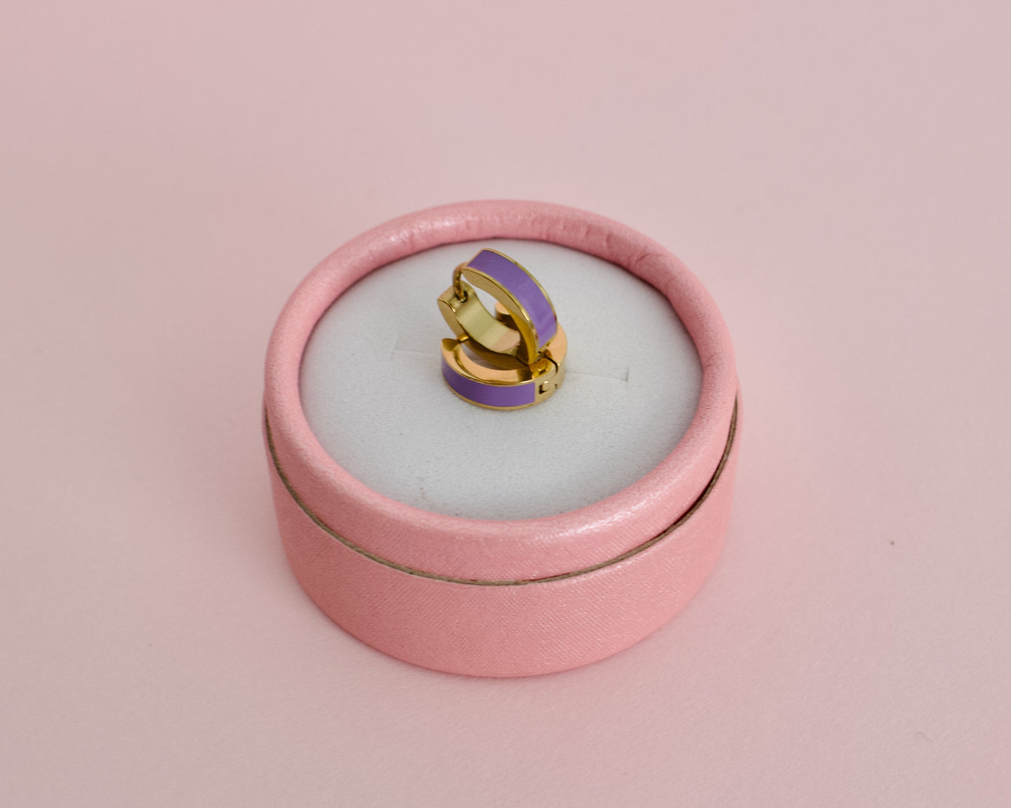 A pair of kids small hypoallergenic gold hoop earrings with a smooth purple enamel inlay, displayed on a pink round box against a solid pale pink background. The earrings are from the brand The Pierced Peanut Earring Co in New Zealand, NZ.