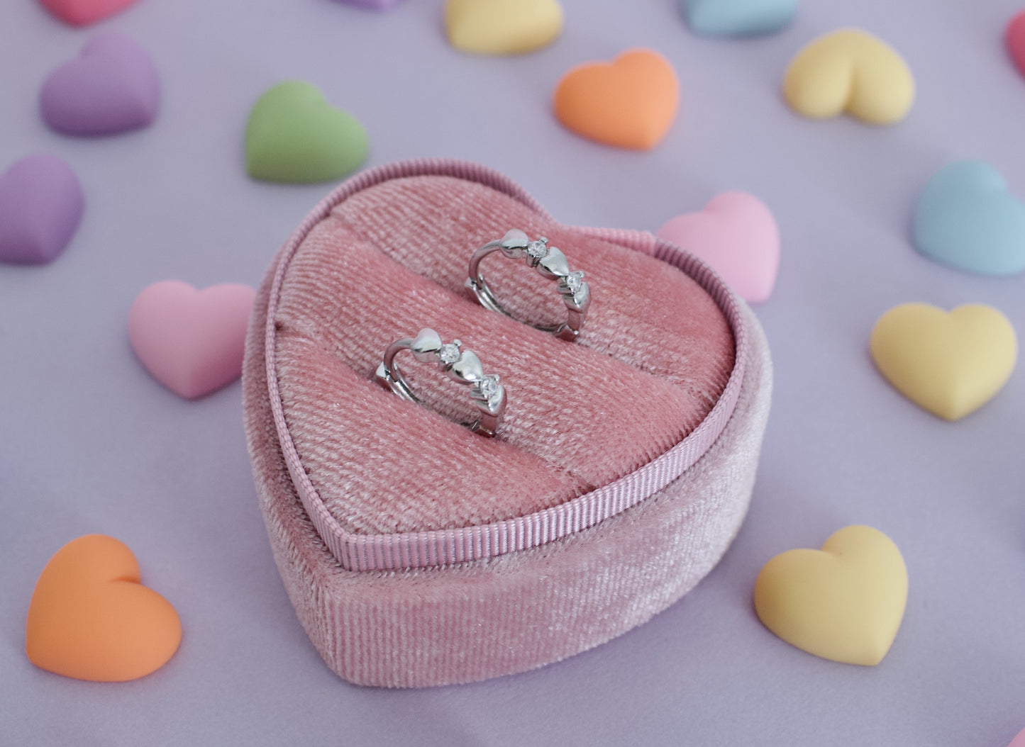 A pair of kids small hypoallergenic silver hoop earrings with heart and diamond detailing, displayed on a pink ring box against a purple background with coloured hearts. The earrings are from the brand The Pierced Peanut Earring Co in New Zealand, NZ.