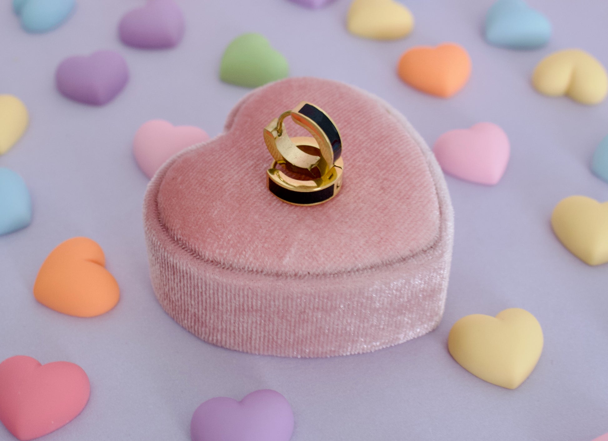 A Pair of kids hypoallergenic huggie hoop earrings with black enamel sections, displayed on top of a pink heart-shaped velvet ring box, surrounded by pastel candy hearts on a lavender background. The earrings are from the brand The Pierced Peanut Earring Co in NZ.
