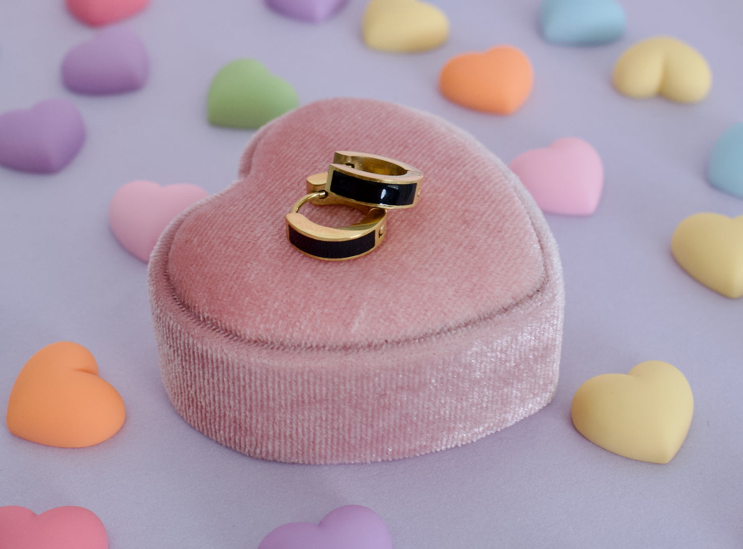 A Pair of kids hypoallergenic huggie hoop earrings with black enamel sections, displayed on top of a pink heart-shaped velvet ring box, surrounded by pastel candy hearts on a lavender background. The earrings are from the brand The Pierced Peanut Earring Co in NZ.