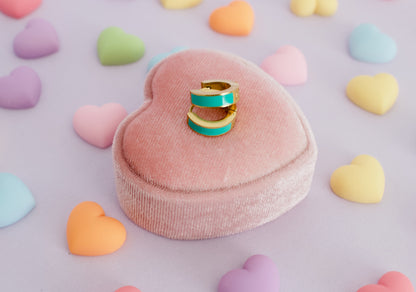 A pair of kids hypoallergenic gold huggie hoop earrings with a turquoise green enamel inlay rests on a pink heart-shaped velvet ring box, surrounded by scattered pastel candy hearts on a lavender background. The earrings are from the brand The Pierced Peanut Earring Co in New Zealand, Nz.