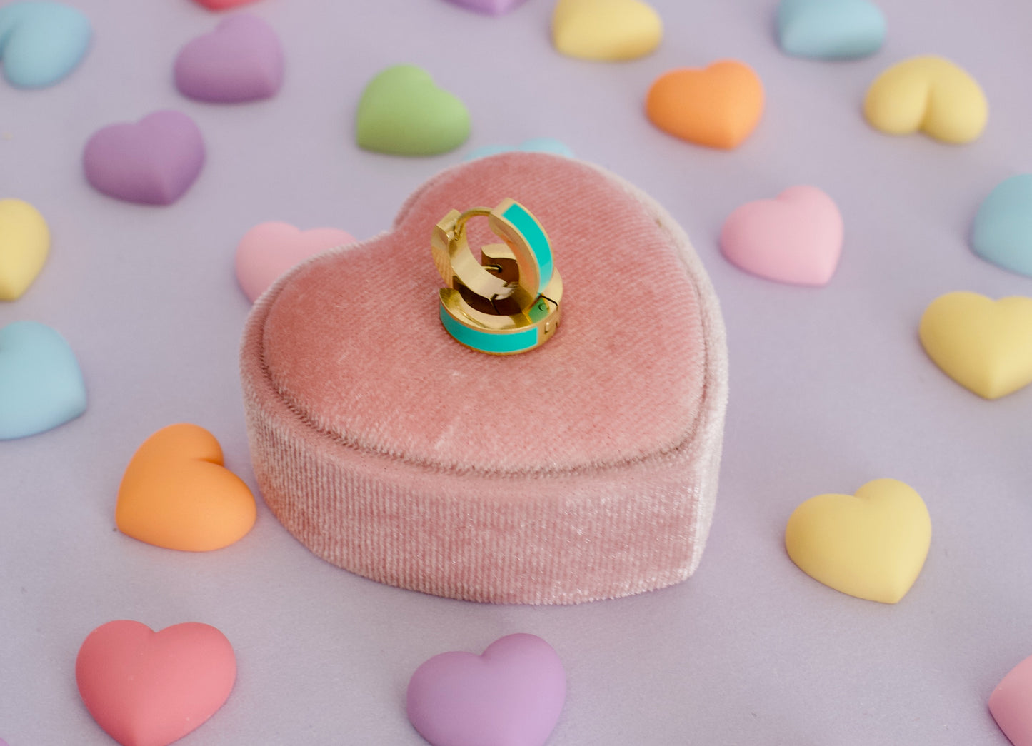 A pair of kids hypoallergenic gold huggie hoop earrings with a turquoise green enamel inlay rests on a pink heart-shaped velvet ring box, surrounded by scattered pastel candy hearts on a lavender background. The earrings are from the brand The Pierced Peanut Earring Co in New Zealand, Nz.