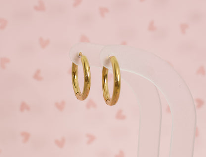 A pair of kids small hypoallergenic gold hoop huggie earrings displayed on a clear acrylic stand against a soft pink background with faint heart patterns. The earrings are from the Pierced Peanut in NZ.