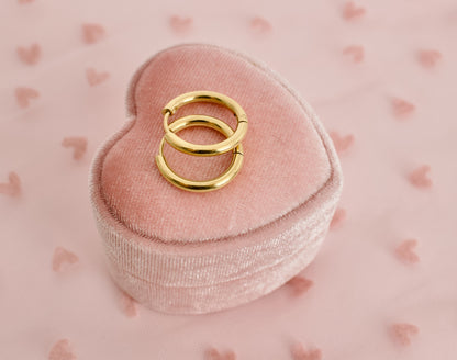 A pair of kids small hypoallergenic gold hoop huggie earrings displayed on a pink heart box against a soft pink background with faint heart patterns. The earrings are from the Pierced Peanut in NZ.