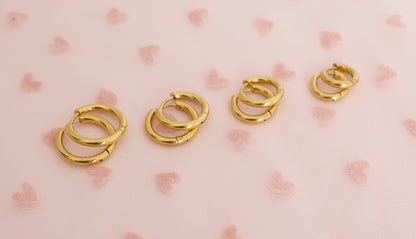 4 pairs of kids small hypoallergenic gold hoop huggie earrings displayed  against a soft pink background with faint heart patterns. The earrings are from the Pierced Peanut in NZ.