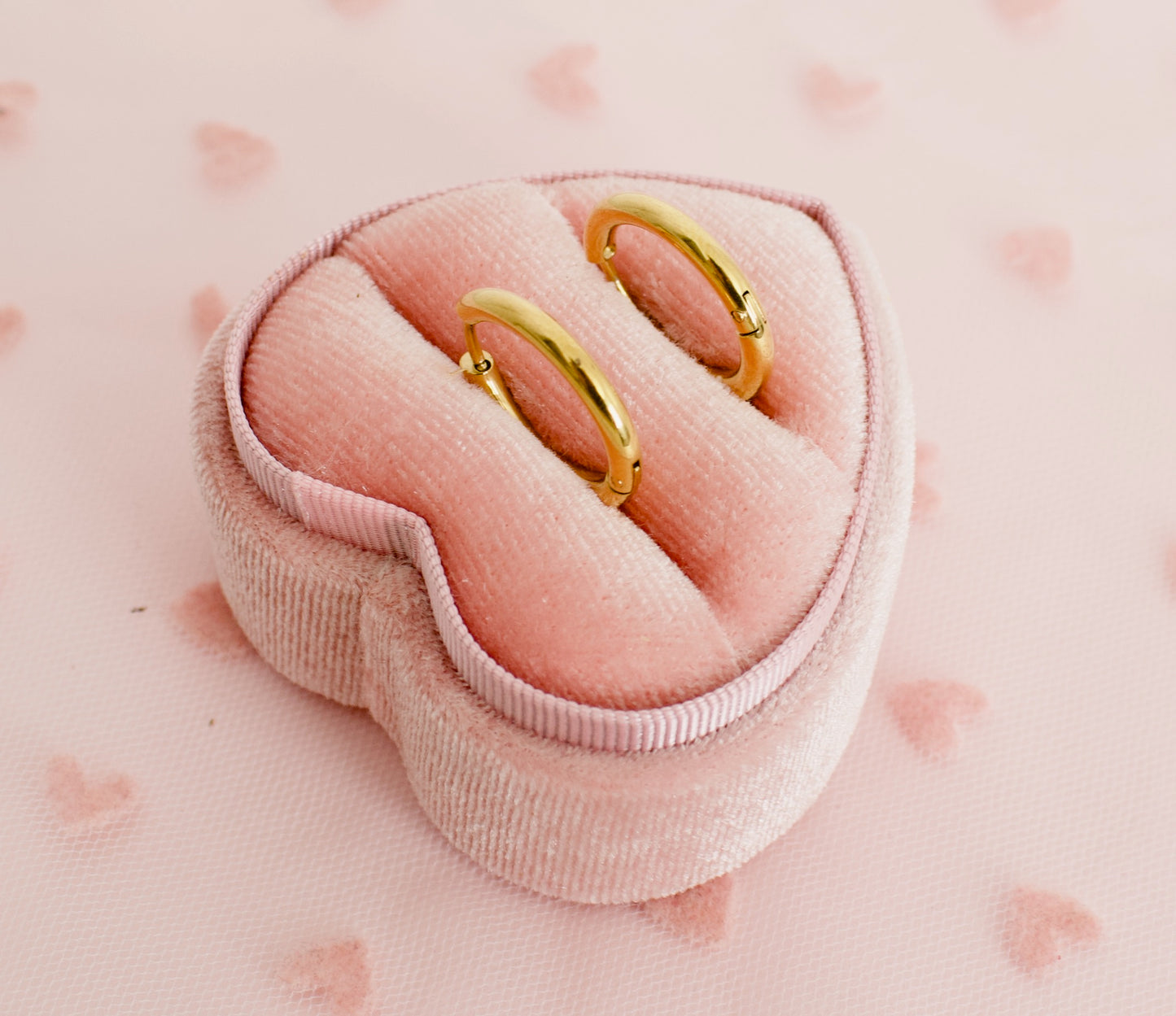 A pair of kids small hypoallergenic gold hoop huggie earrings displayed on a pink heart box against a soft pink background with faint heart patterns. The earrings are from the Pierced Peanut in NZ.
