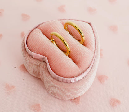 A pair of kids small hypoallergenic gold hoop huggie earrings displayed on a pink heart box against a soft pink background with faint heart patterns. The earrings are from the Pierced Peanut in NZ.