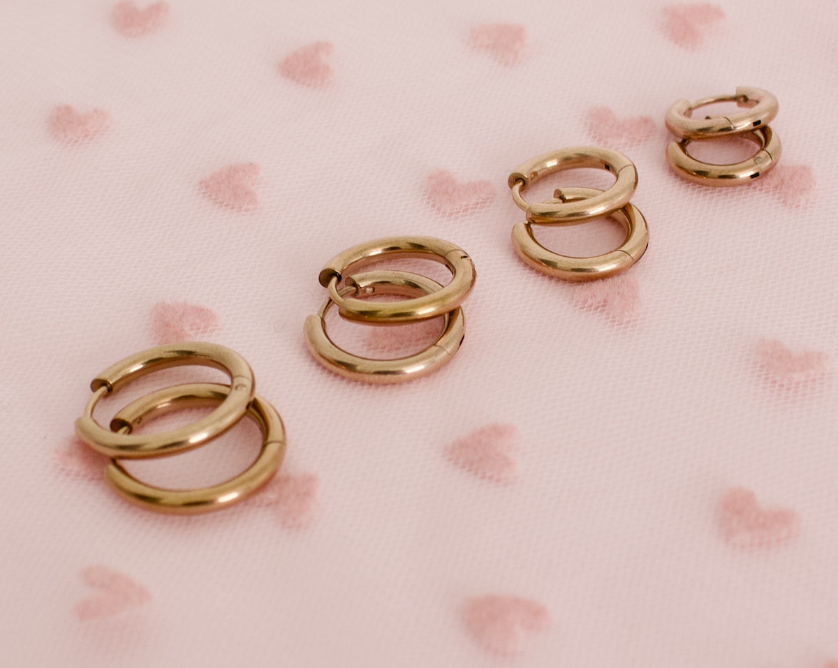 Four pairs of hypoallergenic kids rose gold-toned huggie hoop earrings arranged on a light pink fabric background with a subtle heart pattern. Earrings are from the brand the Pierced Peanut earring co in New Zealand.