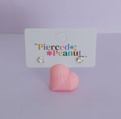 A pair of kids small hypoallergenic heart-shaped stud earrings with clear rhinestones in gold settings, displayed on a white branded card that reads 'The Pierced * Peanut * Design Co.' A pink heart-shaped stand sits below the card on a soft lavender studio background.