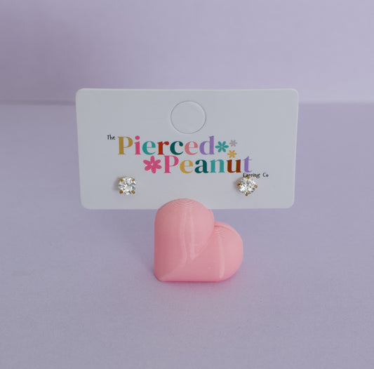 A pair of kids small hypoallergenic heart-shaped stud earrings with clear rhinestones in gold settings, displayed on a white branded card that reads 'The Pierced * Peanut * Design Co.' A pink heart-shaped stand sits below the card on a soft lavender studio background.