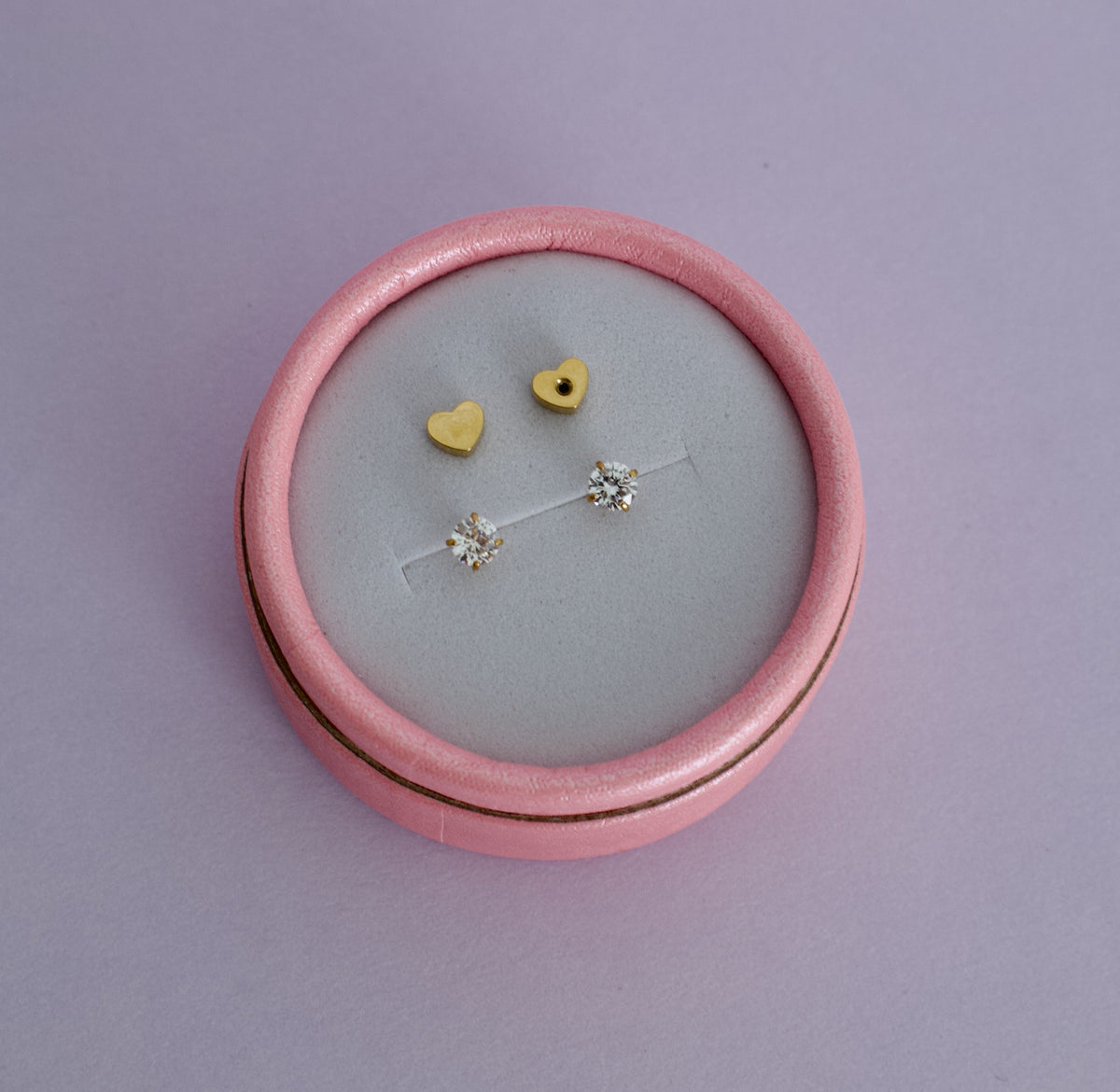 A pink round jewelry box with a light gray velvet insert, holding a pair of kida hypoallergenic gold diamond stud earrings with heart shaped screwbacks. Earrings are from the Pierced Peanut in Nz