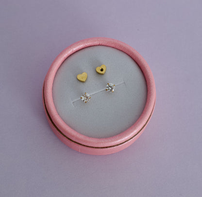 A pink round jewelry box with a light gray velvet insert, holding a pair of kida hypoallergenic gold diamond stud earrings with heart shaped screwbacks. Earrings are from the Pierced Peanut in Nz