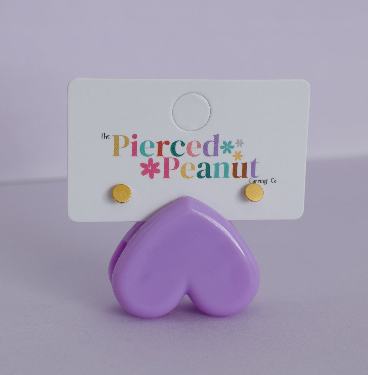 A pair of kids small hypoallergenic gold stud earrings attached to a white display card, with a glossy lavender heart-shaped stand in front. The card reads 'The Pierced Peanut Earring Co'.