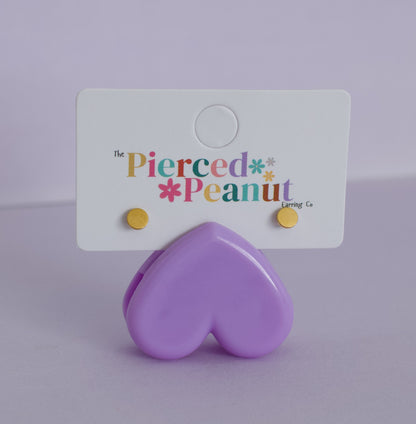 A pair of kids small hypoallergenic gold stud earrings attached to a white display card, with a glossy lavender heart-shaped stand in front. The card reads 'The Pierced Peanut Earring Co'.