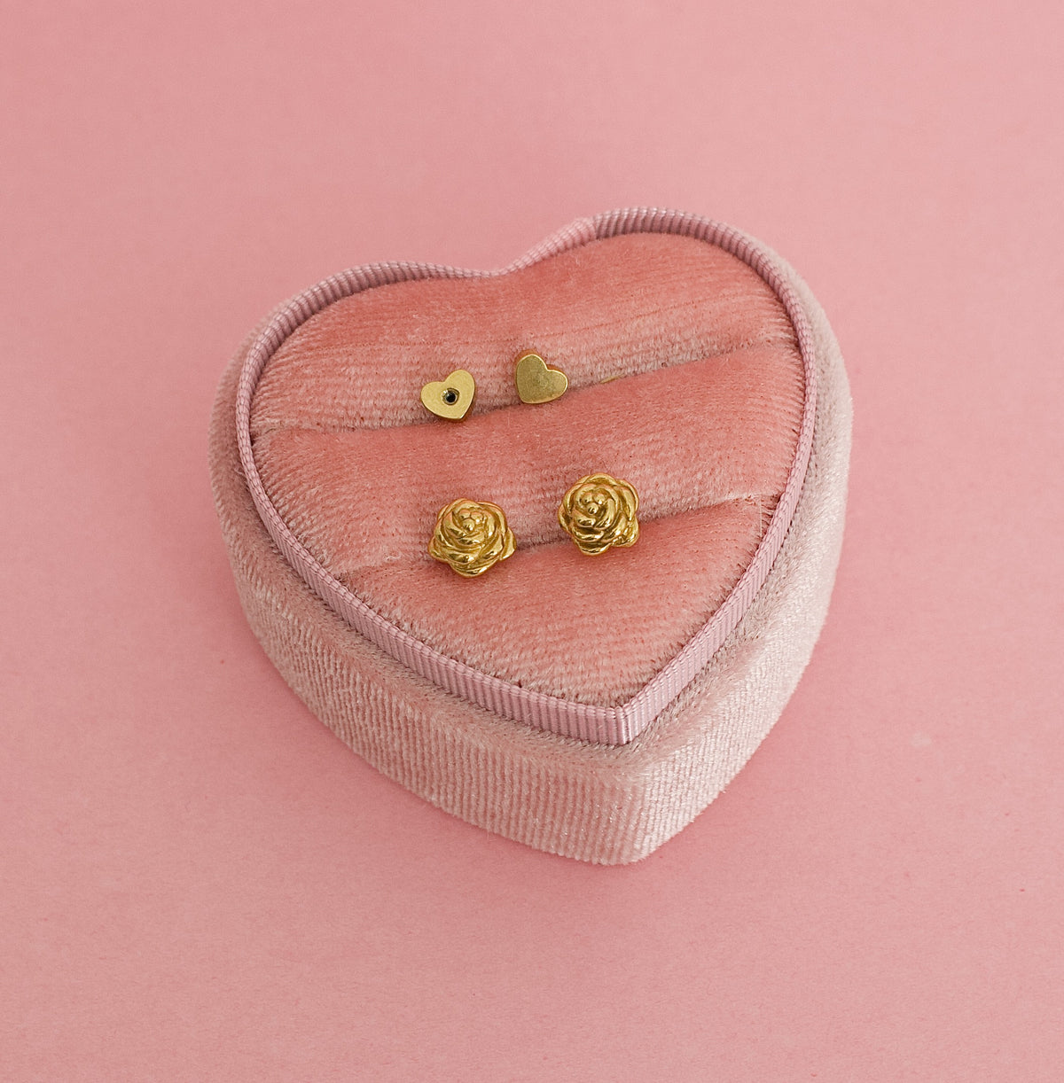 A pair of kids hypoallergenic gold earrings shaped like rose flowers, displayed on a pink heart shaped box. The earrings are screwback studs from the Pierced Peanut in Nz.