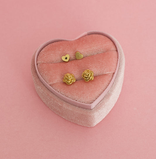 A pair of kids hypoallergenic gold earrings shaped like rose flowers, displayed on a pink heart shaped box. The earrings are screwback studs from the Pierced Peanut in Nz.