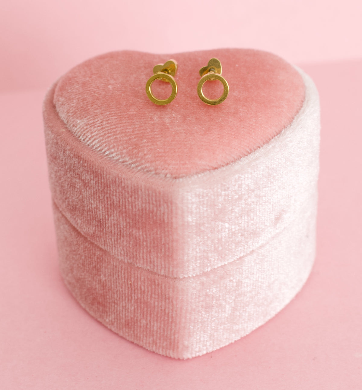 A pair of small gold halo earrings displayed on top of a pink velvet heart-shaped jewelry box against a solid pink background. Earrings are kids hypoallergenic gold screwback studs from the Pierced Peanut earring co in Nz