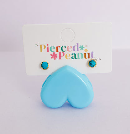 A pair of gold-framed stud earrings with round turquoise blue cabochon stones, displayed on a white backing card that reads 'The Pierced Peanut Earring Co.' with a light blue heart-shaped prop in front.