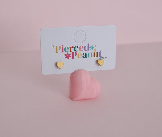 A pair of kids small hypoallergenic gold heart stud earrings displayed on a white branded card stand, with a pink heart-shaped prop below on a soft pink background. The card reads 'the Pierced Peanut Earring Co'.