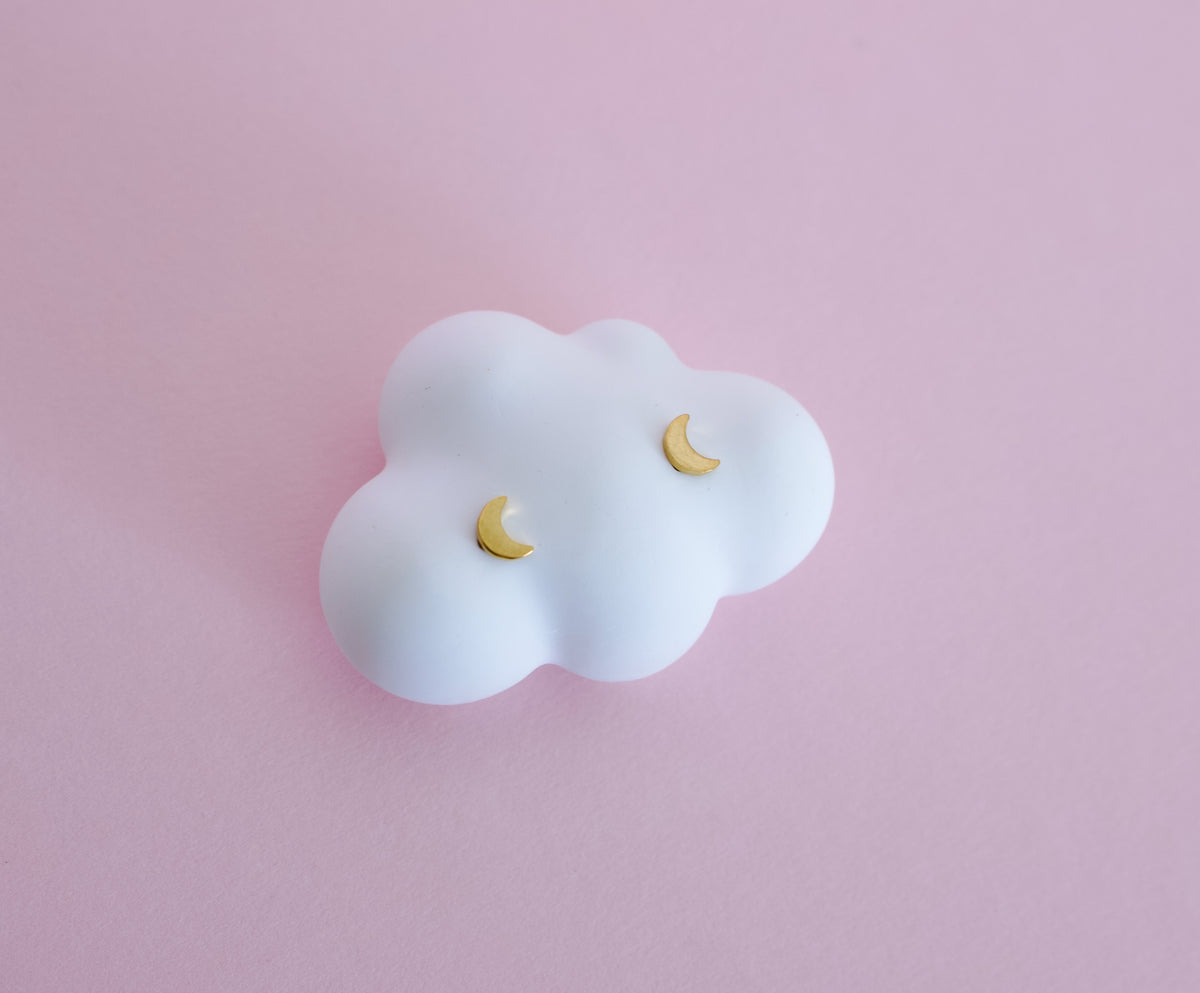 A pair of small gold crescent moon stud earrings displayed on a white cloud-shaped jewellery stand against a soft pink background. Earrings are for kids, made of hypoallergenic materials and have heart shaped screwbacks for the stud, from the Pierced Peanut Earring Co in Nz