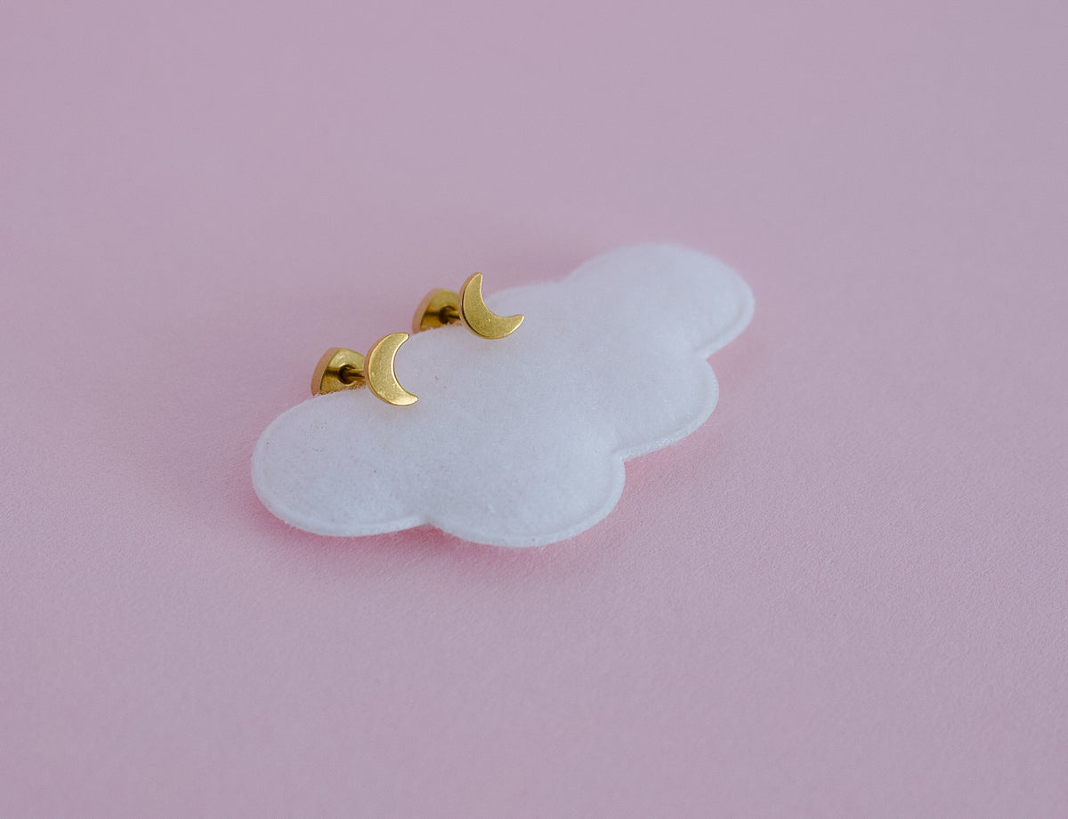 A pair of kids small hypoallergenic gold crescent moon screwback stud earrings displayed on a white cloud-shaped stand against a soft pink background. Earrings are from the Pierced Peanut in Nz
