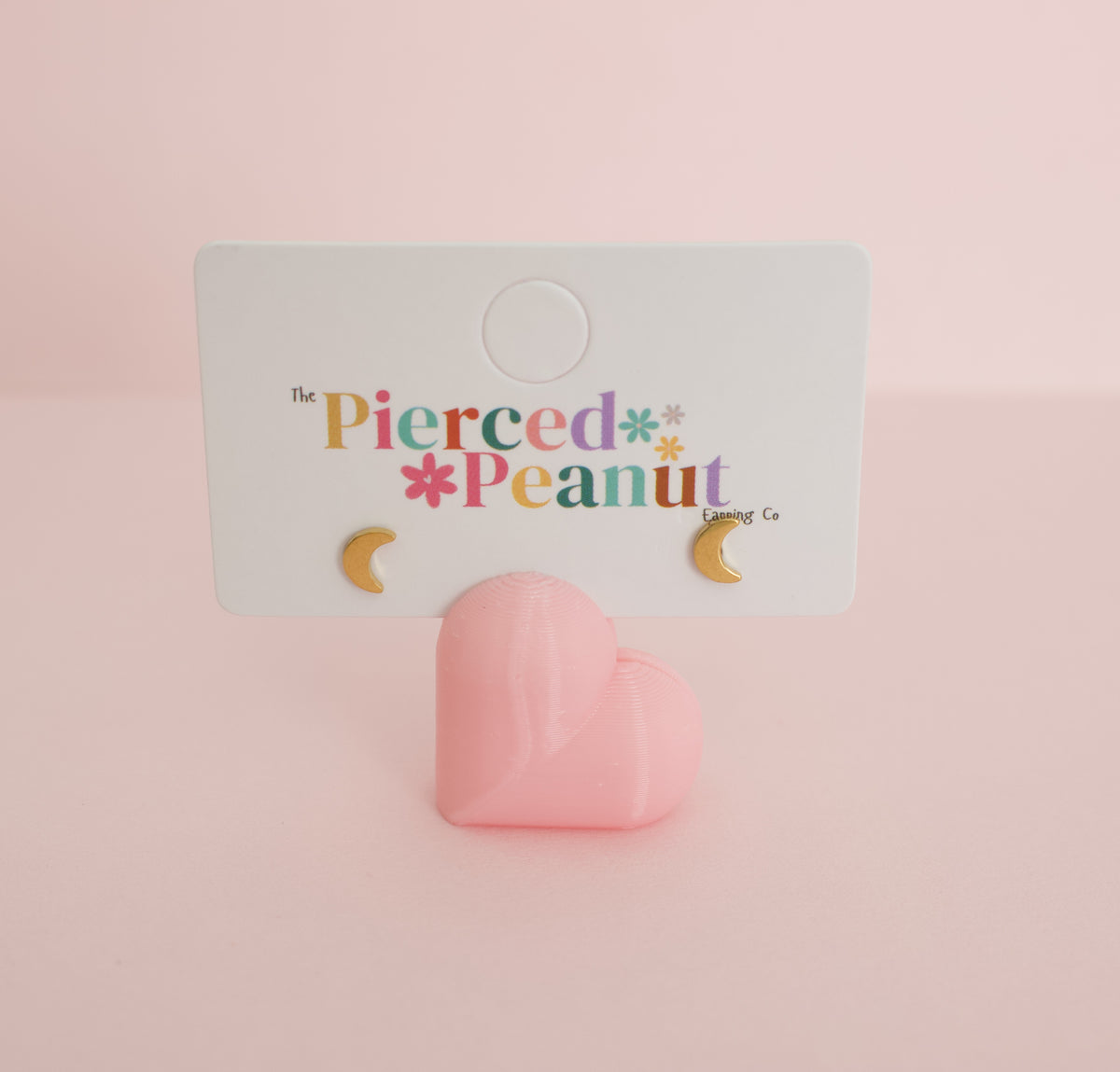 A pair of kids small hypoallergenic gold crescent moon screwback stud earrings displayed on a white branded card holder against a soft pink background. The card reads 'The Pierced Peanut Earring Co.