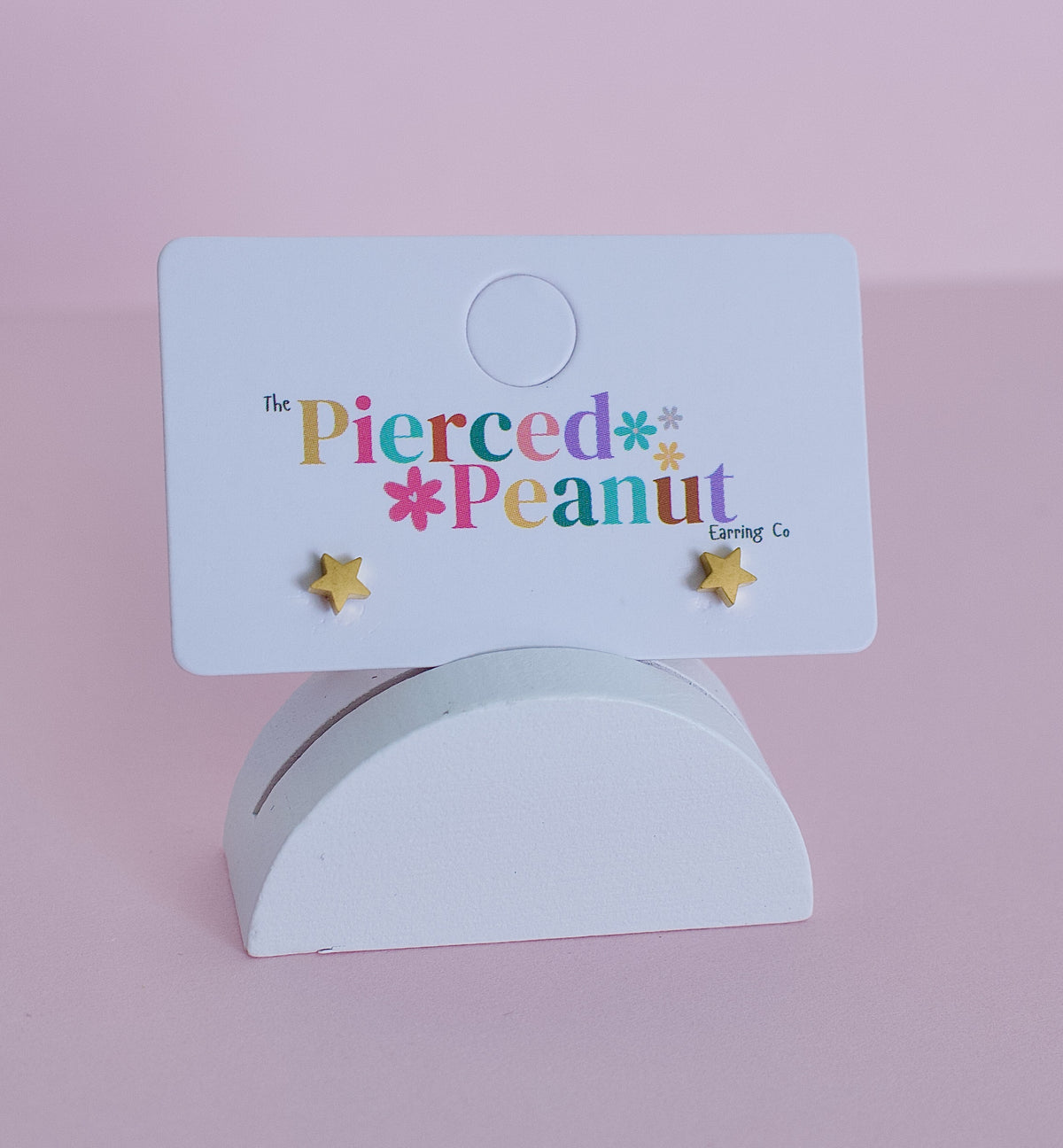 Two small kids gold hypoallergenic star screwback stud earrings displayed on a white card from 'The Pierced Peanut Earring Co' against a soft pink background.