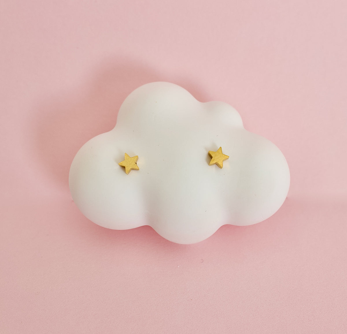 A pair of kids small hypoallergenic gold star screwback stud earrings displayed on a white cloud-shaped jewellery stand against a soft pink background. Earrings are from the Pierced Peanut in New Zealand.