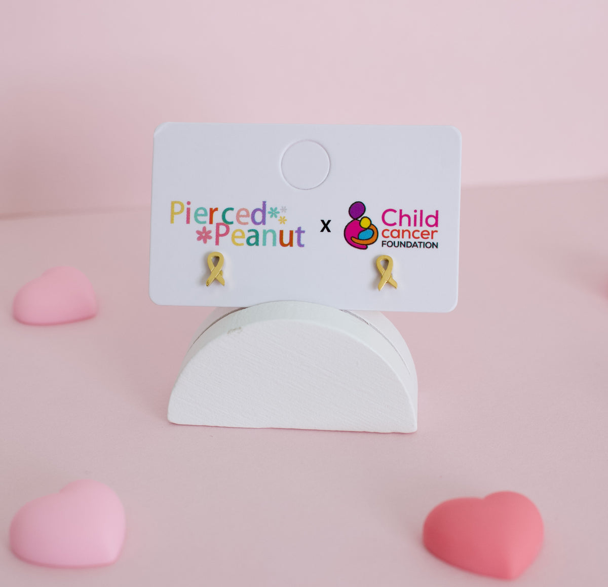 A pair of small hypoallergenic yellow ribbon screwback stud earrings displayed on a white backing card that reads 'Pierced Peanut x Child Cancer Foundation,' set on a white stand against a soft pink background with pink heart props.