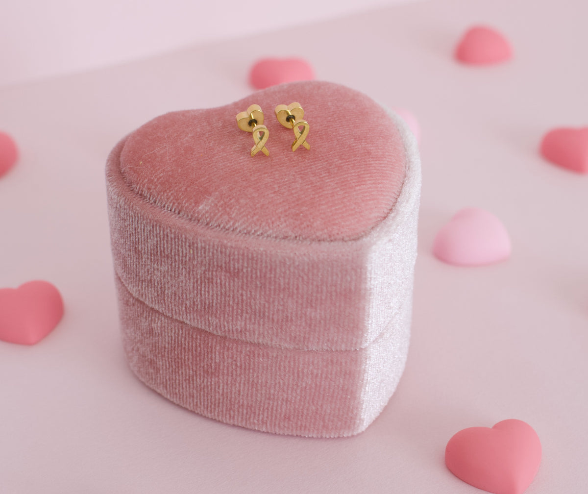 A pair of small hypoallergenic gold heart-shaped screwback stud earrings displayed on top of a pink velvet heart-shaped jewelry box, with pink heart-shaped props scattered on a soft pink background. The earrings are from the Pierced Peanut in collaboration with the Child Cancer Foundation, Nz.