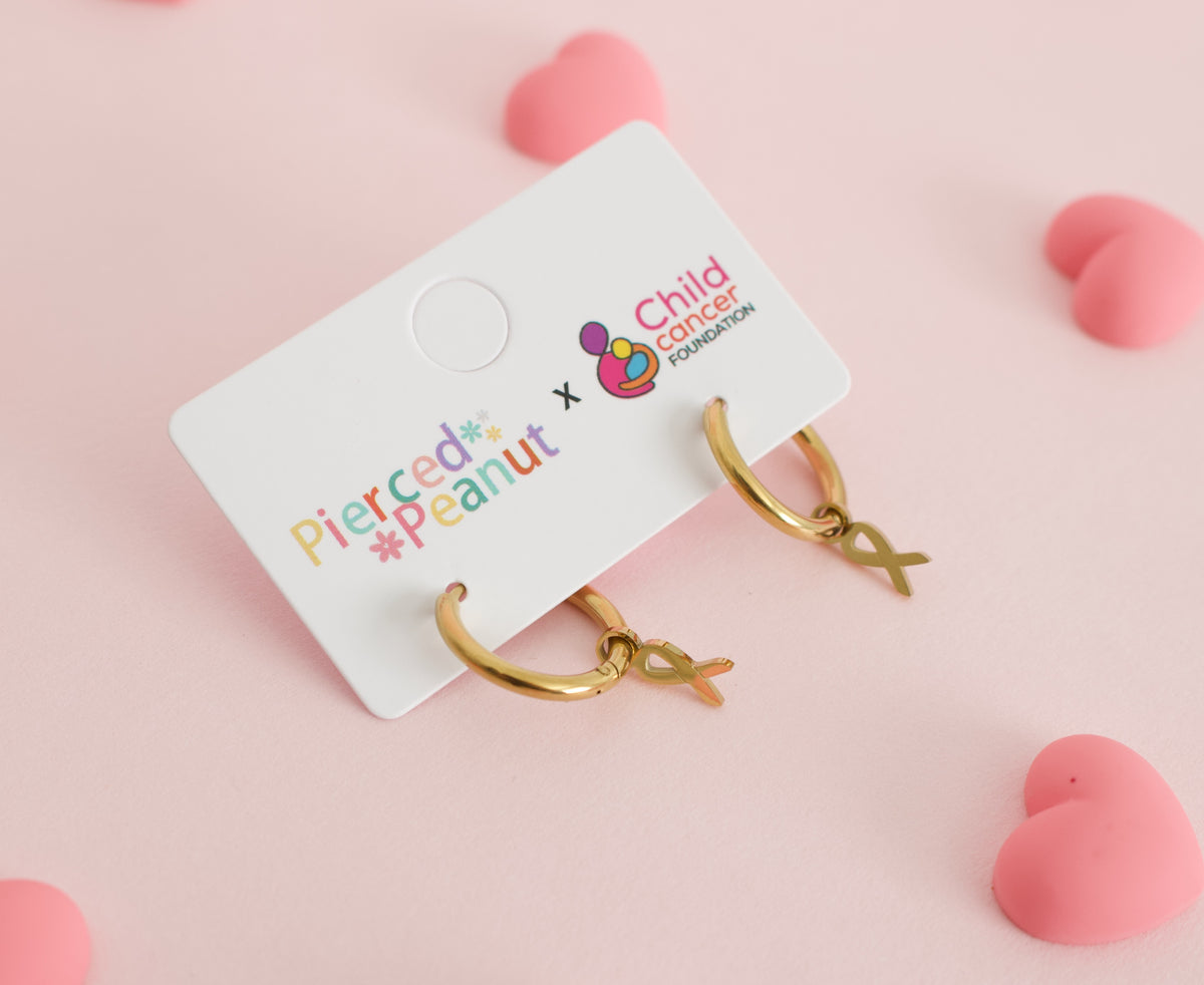 A pair of hypoallergenic gold huggie hoop earrings with gold ribbon charms on a branded white card from the Pierced Peanut and Child Cancer Foundation Nz