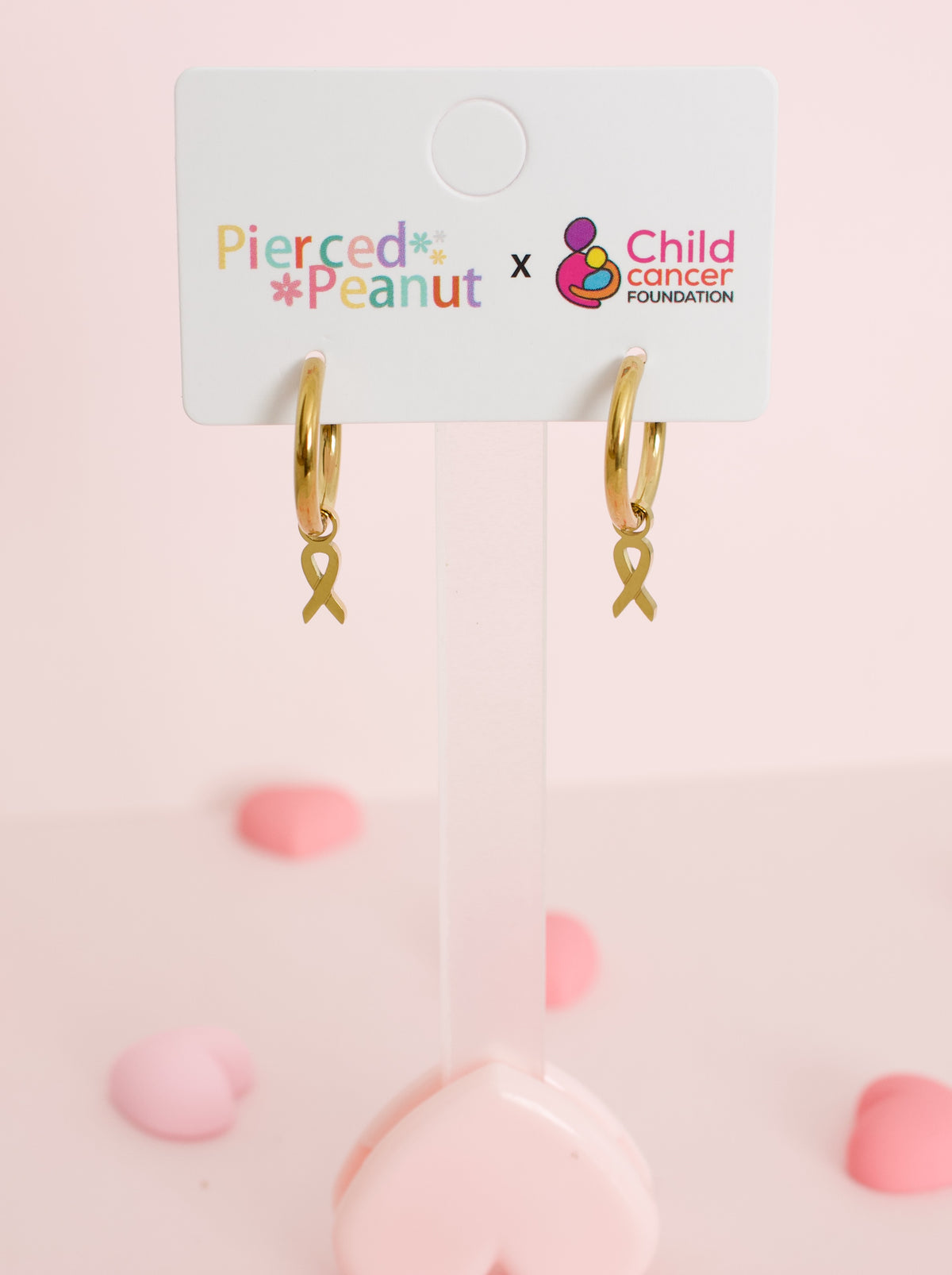 A pair of hypoallergenic gold huggie hoop earrings with gold ribbon charms on a branded white card from the Pierced Peanut and Child Cancer Foundation Nz