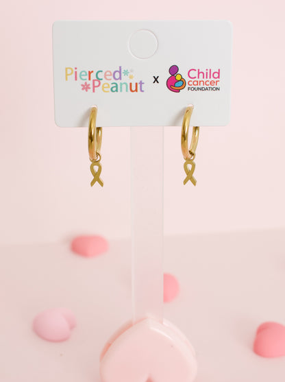 A pair of hypoallergenic gold huggie hoop earrings with gold ribbon charms on a branded white card from the Pierced Peanut and Child Cancer Foundation Nz