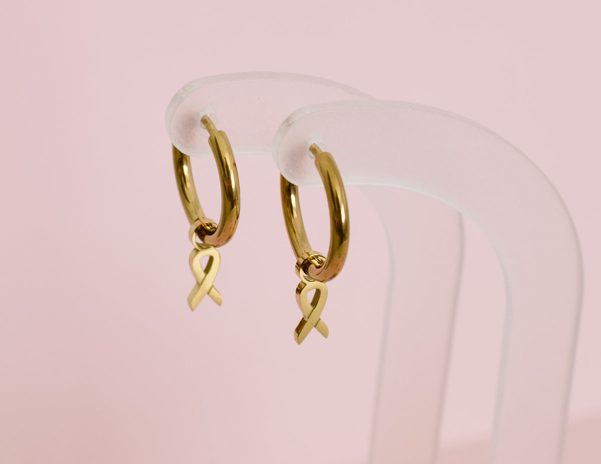 A pair of hypoallergenic gold huggie hoop earrings with gold ribbon charms from the Pierced Peanut in collaboration with the  Child Cancer Foundation Nz
