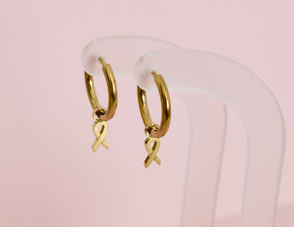A pair of hypoallergenic gold huggie hoop earrings with gold ribbon charms from the Pierced Peanut in collaboration with the  Child Cancer Foundation Nz
