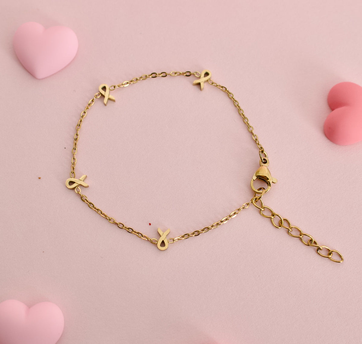 A gold chain bracelet with four small gold ribbon charms, displayed on a soft pink background with decorative pink heart shapes. The bracelet is made by the Pierced Peanut in collaboration with the Child Cancer Foundation Nz as a fundraiser.