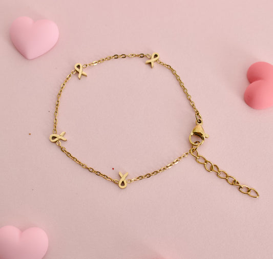A gold chain bracelet with four small gold ribbon charms, displayed on a soft pink background with decorative pink heart shapes. The bracelet is made by the Pierced Peanut in collaboration with the Child Cancer Foundation Nz as a fundraiser.
