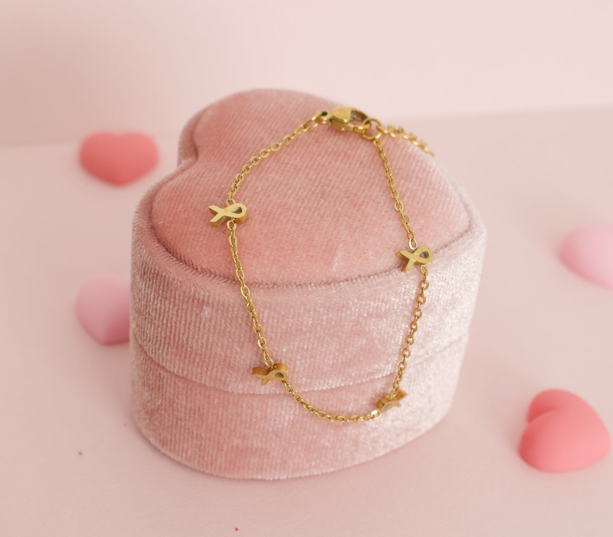 A delicate gold chain bracelet with small gold ribbon charms, displayed on a pink velvet heart-shaped jewelry box against a soft pink background with decorative heart props. 
The bracelet is made by the Pierced Peanut in collaboration with the Child Cancer Foundation Nz as a fundraiser.
