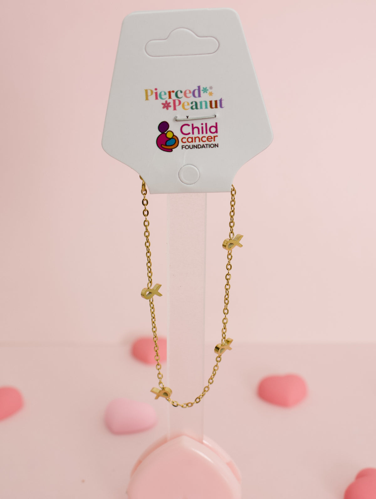 A gold chain bracelet with small gold ribbon charms, displayed on a white Pierced Peanut Child Cancer Foundation backing card against a soft pink background with pink heart-shaped props.