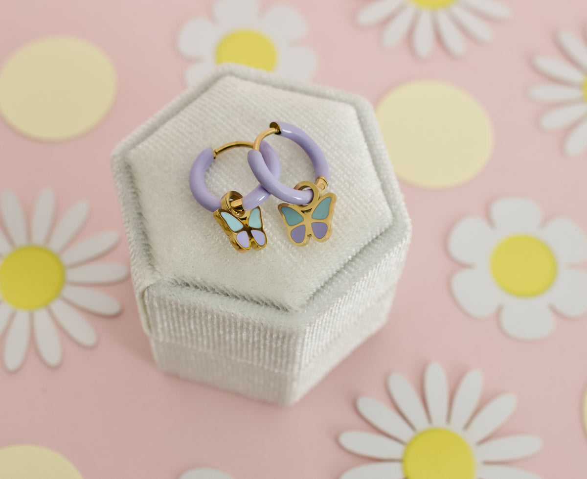 A pair of kids small hypoallergenic butterfly hoop earrings with lavender purple hoops and gold-plated butterfly charms featuring mint green and lavender enamel, displayed on a white box against a soft pink background with flowers. Earrings are from the Pierced Peanut Earring co in Nz